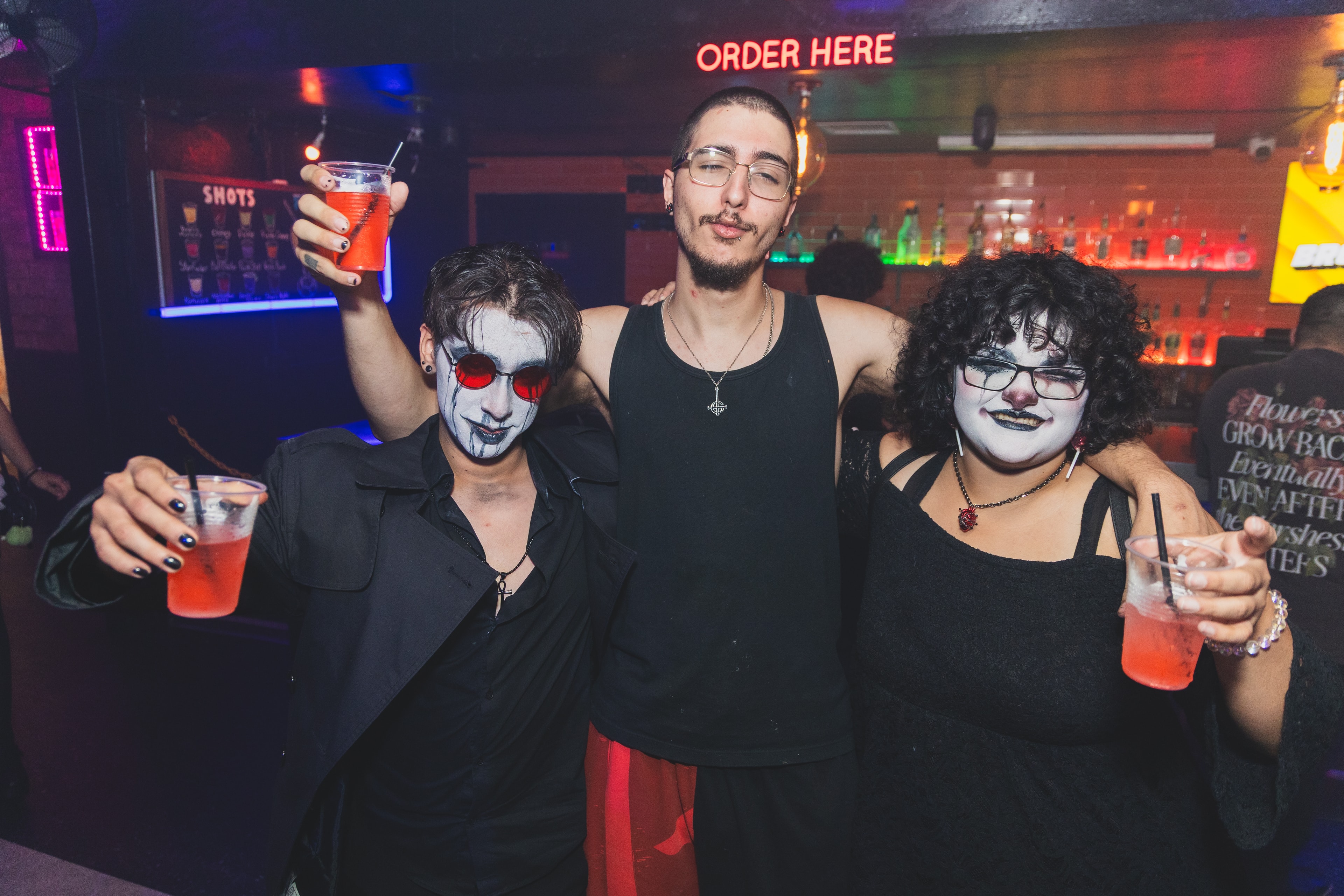 Three friends in goth face paint raising drinks at the bar at SATX Emo Club