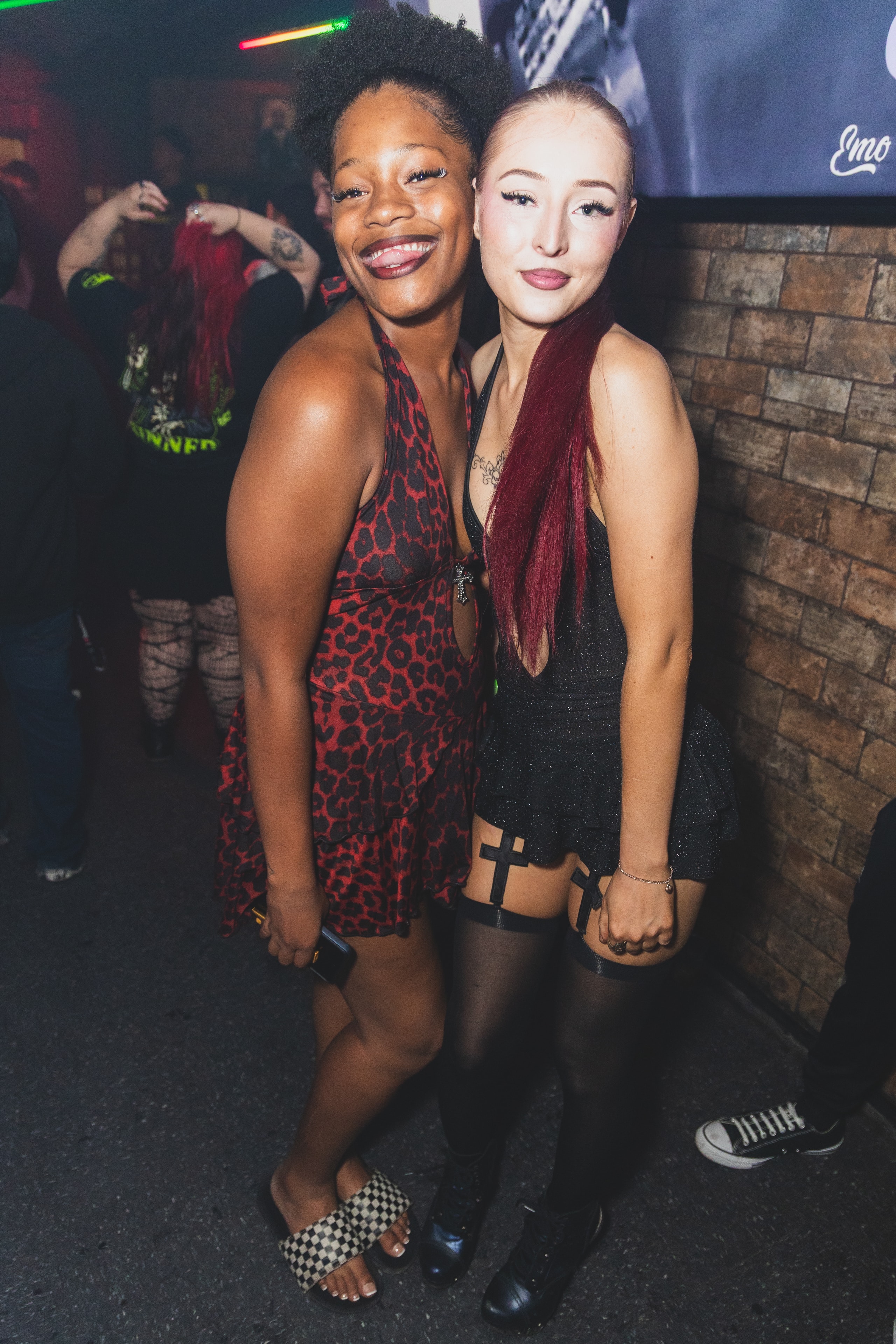 Two women posing together at SATX Emo Club with Emo Night screen in background
