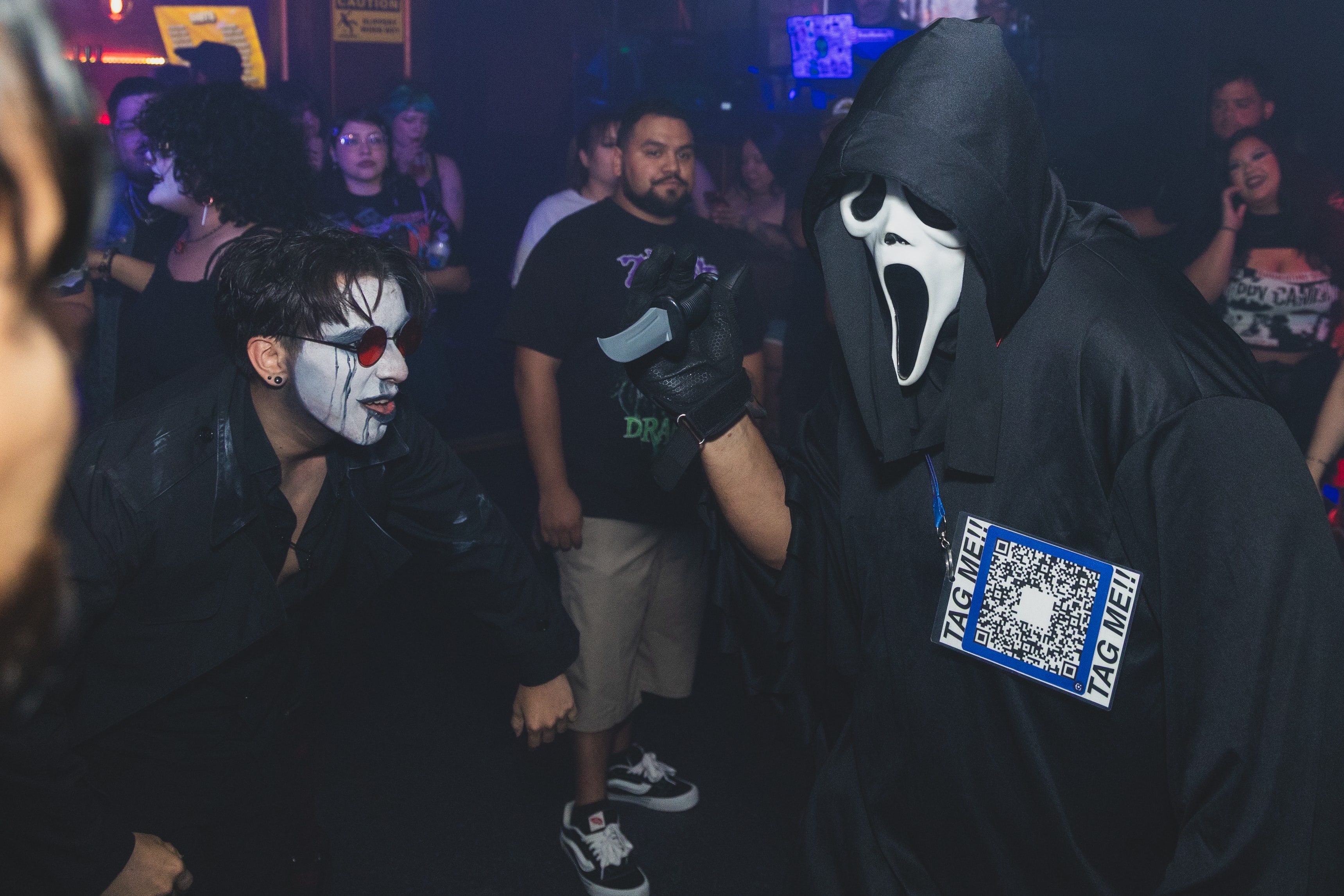 Ghostface with TAG ME QR code lanyard facing off with goth face paint attendee at SATX Emo Club