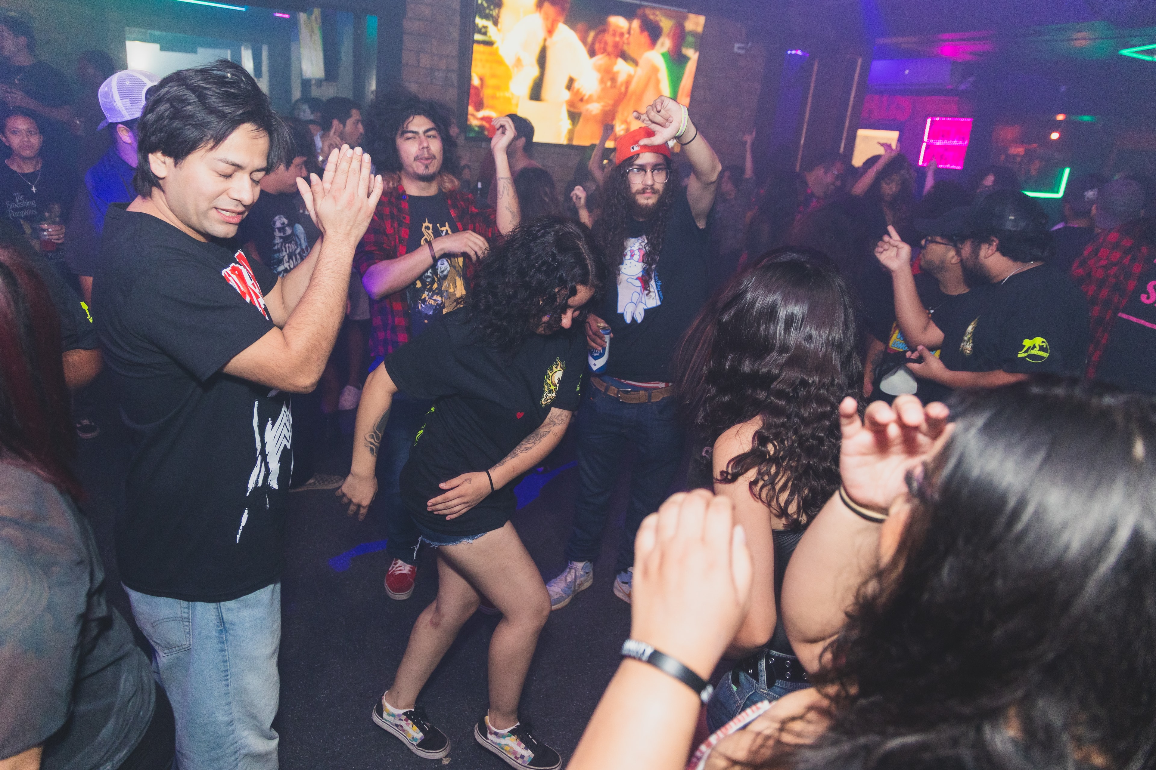 Packed dance floor with band tees and red flannel under neon lights at SATX Emo Club